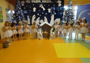 Dziewczynki przebrane za aniołki stoją wokół szopki ze złożonymi rączkami. W szopce siedzi dziewczynka przebrana za Maryję i chłopiec przebrany za Józefa.